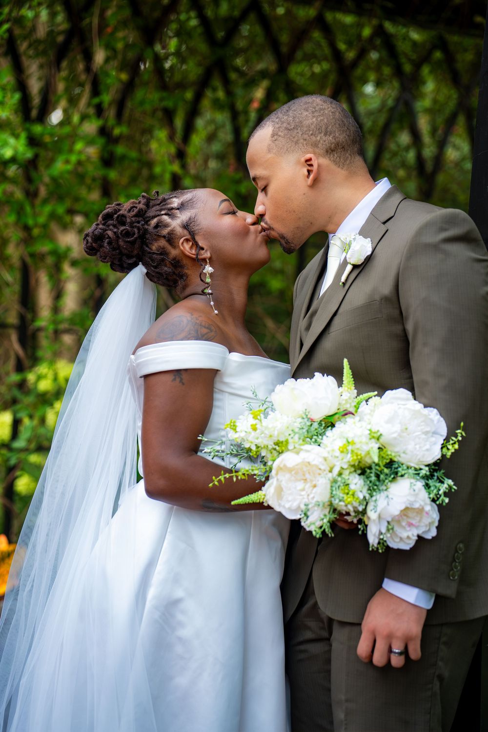 A bride and groom share a kiss outdoors, with the bride holding a white peony bouquet on their wedding day.