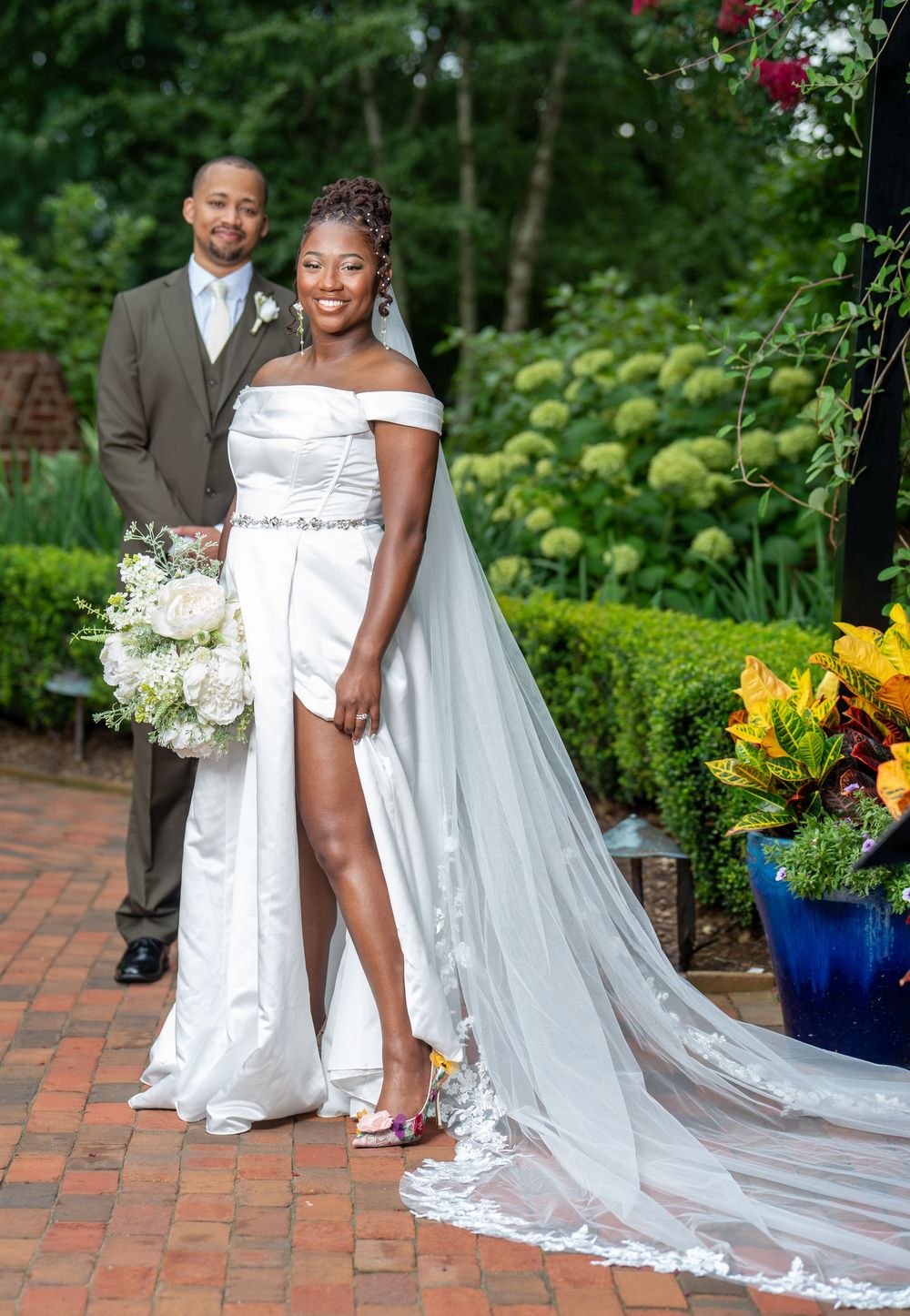 A bride in a white off-shoulder gown with a long veil poses with her groom in a garden setting.