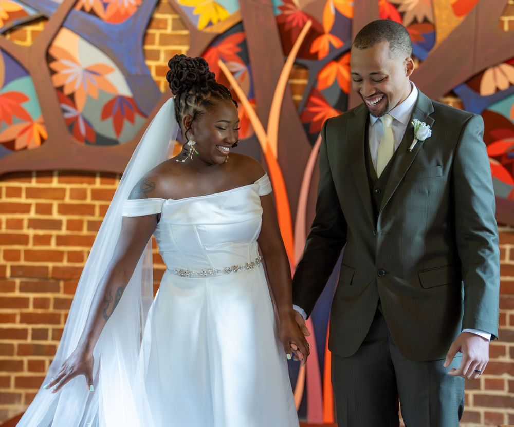 A joyful bride and groom walk hand in hand, laughing together in front of a colorful mosaic wall.