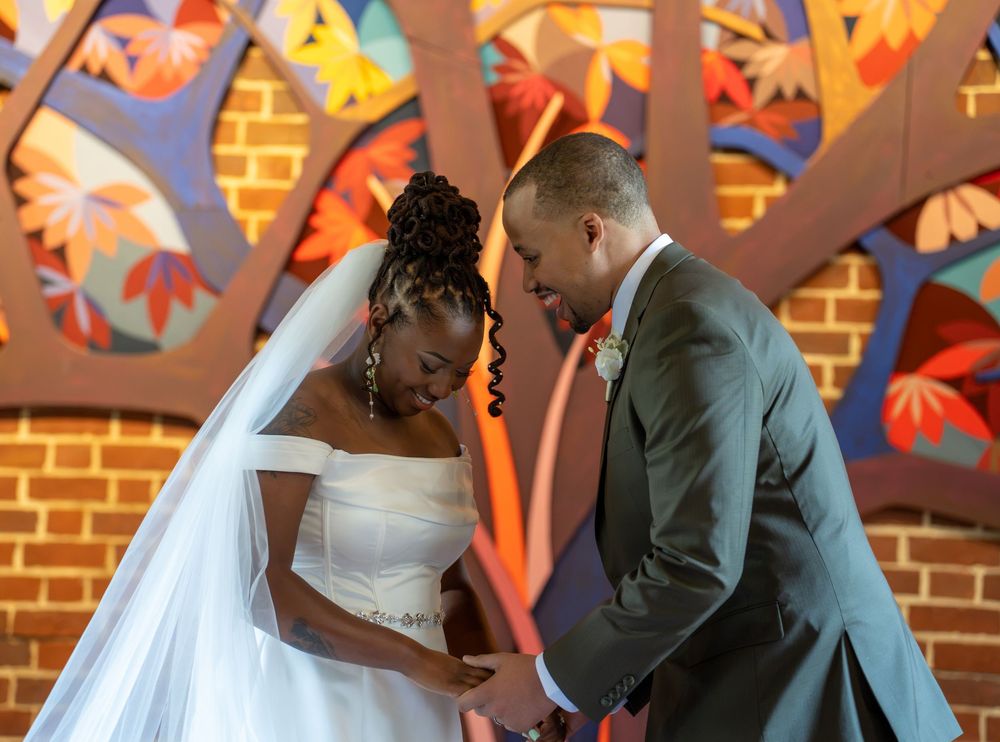 A bride and groom bow their heads together in prayer before a colorful stained glass window during their ceremony.
