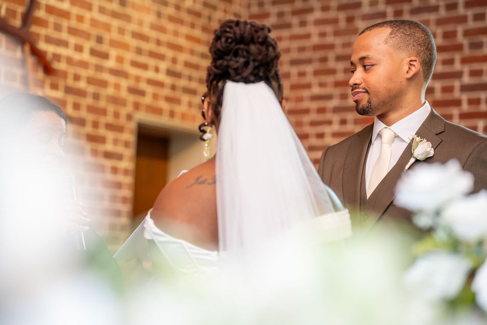A groom gazes lovingly at his bride during their wedding ceremony inside a brick church with floral decorations.