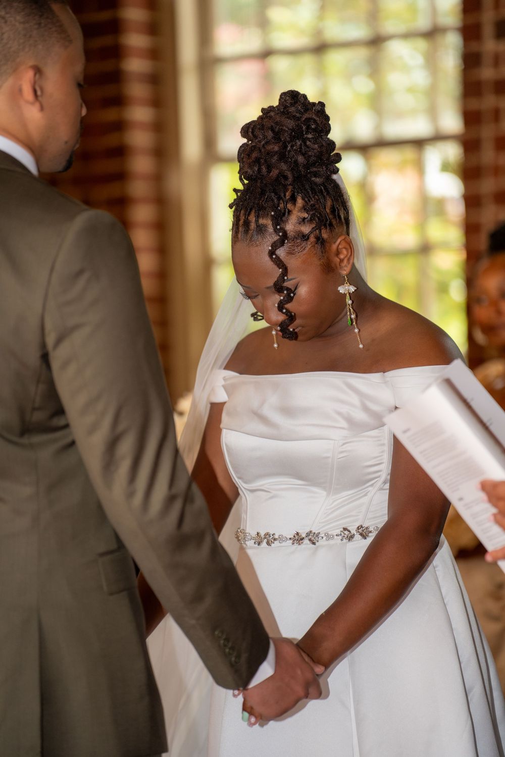A bride in an off-shoulder white gown holds hands with her groom while reading vows inside a brick chapel.