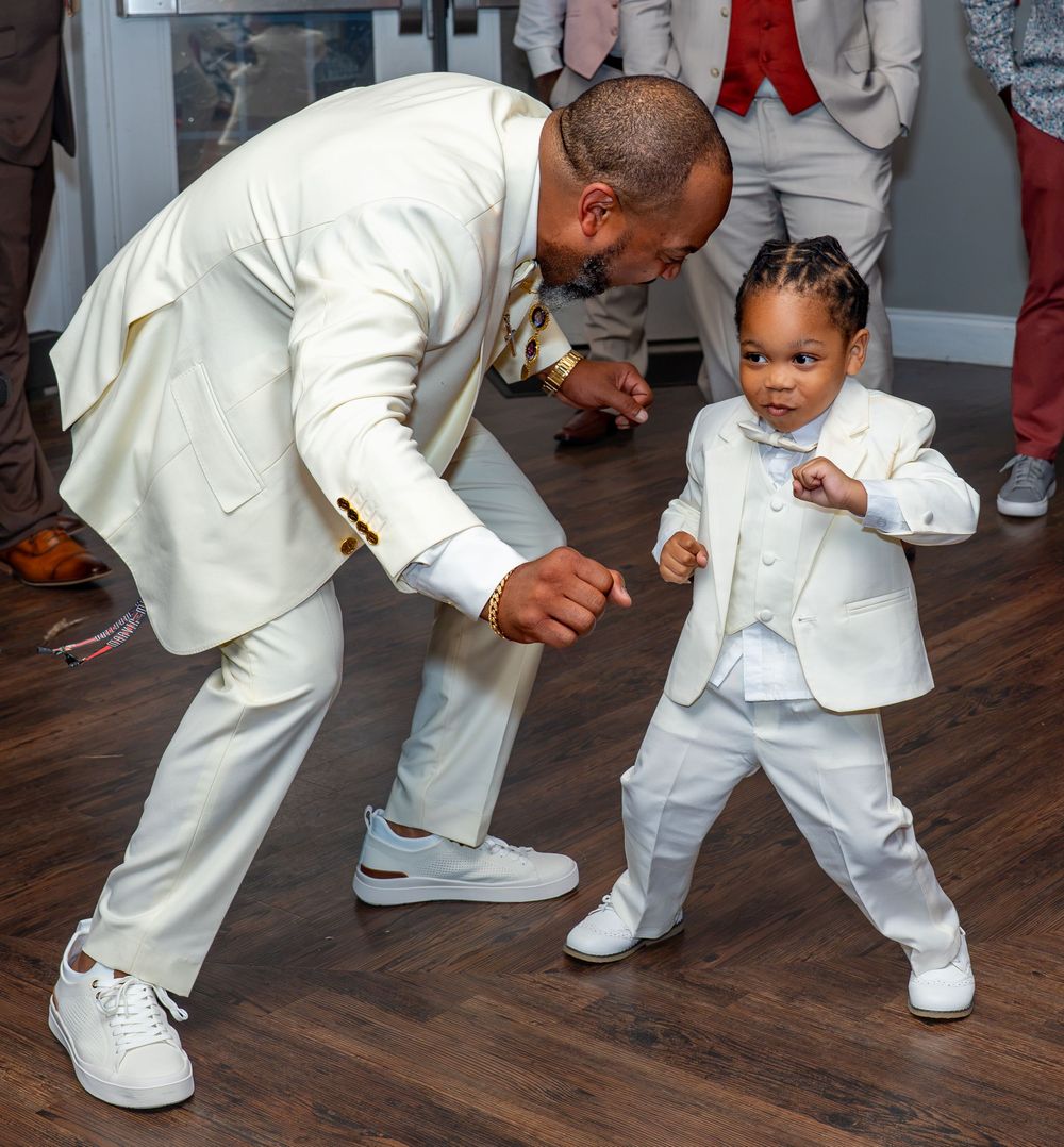 A man in a white suit dances playfully with a toddler dressed in a matching white outfit at a celebration.
