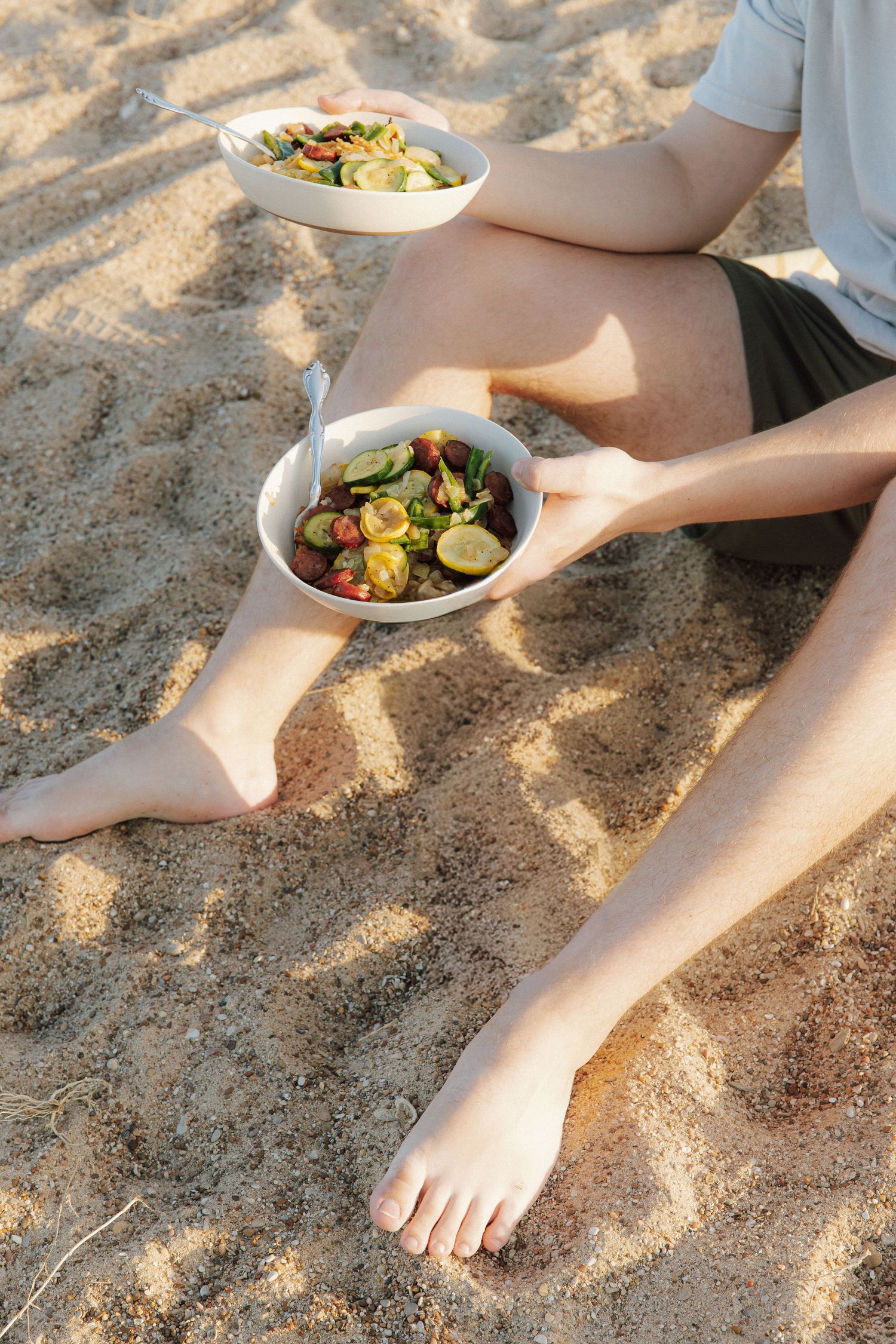 Person sitting on sandy beach holding two bowls of fresh vegetable salad with cucumbers and tomatoes.