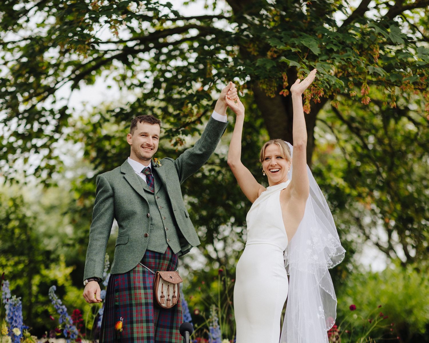 Joyful bride and kilt-wearing groom raise hands in celebration beneath a leafy tree.