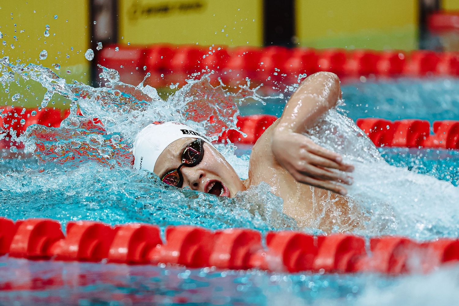 Competitive swimmer in white cap races freestyle stroke in pool with red lane dividers during championship event.