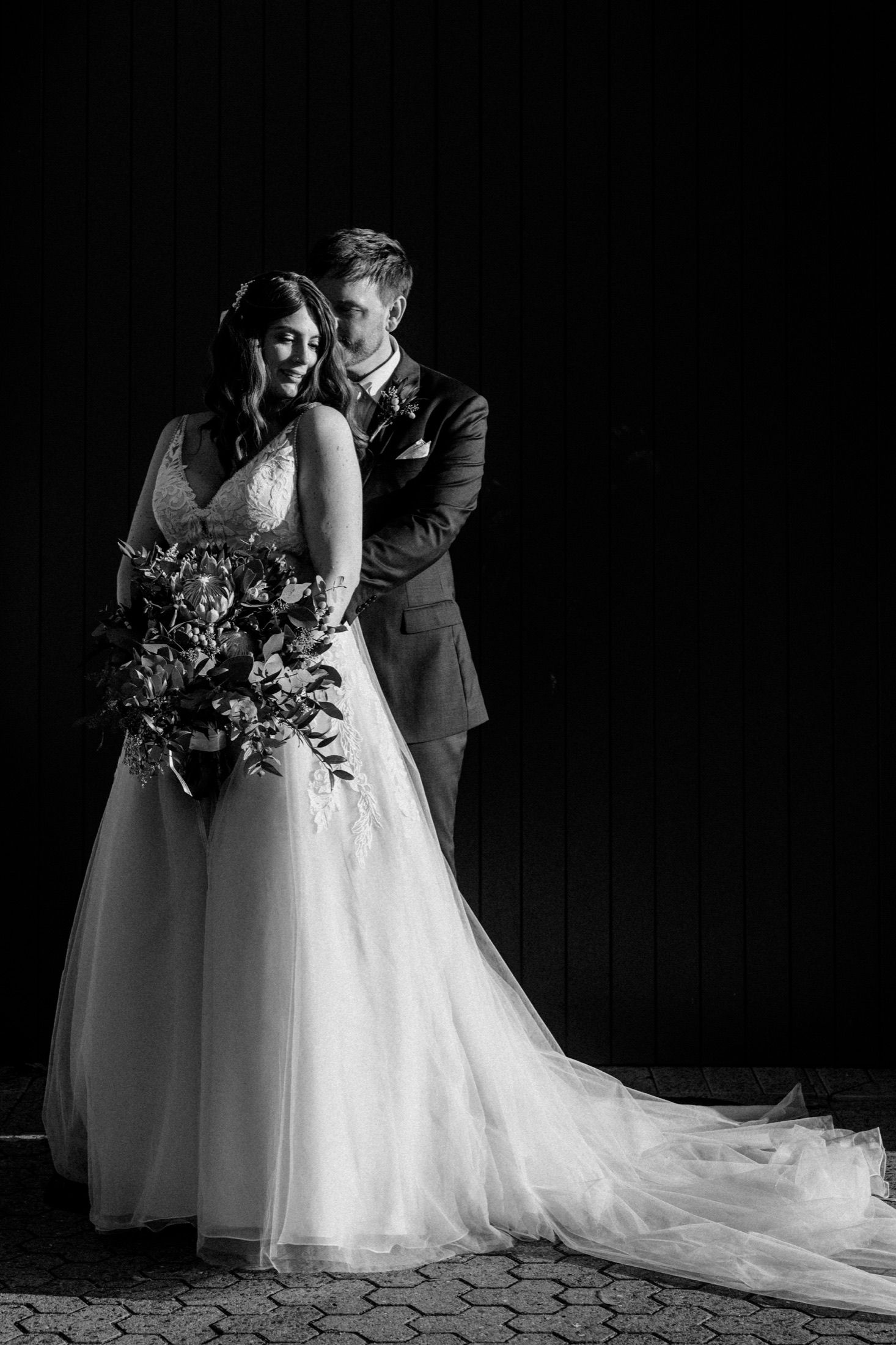 Black and white wedding portrait of bride in flowing gown holding bouquet, groom in suit embracing her from behind.