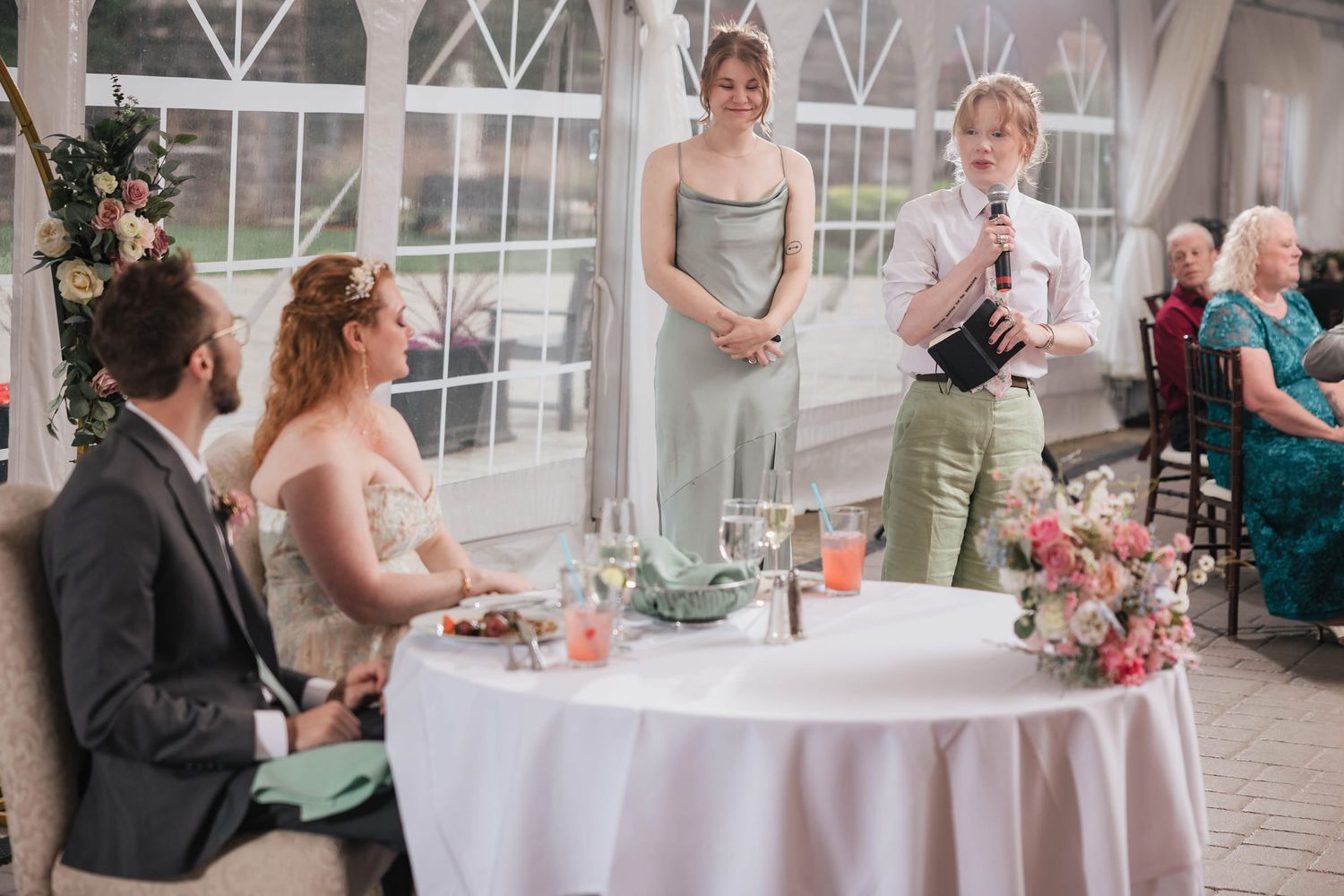 Maid of honor giving a speech from notes, a second bridesmaid standing beside her listening.