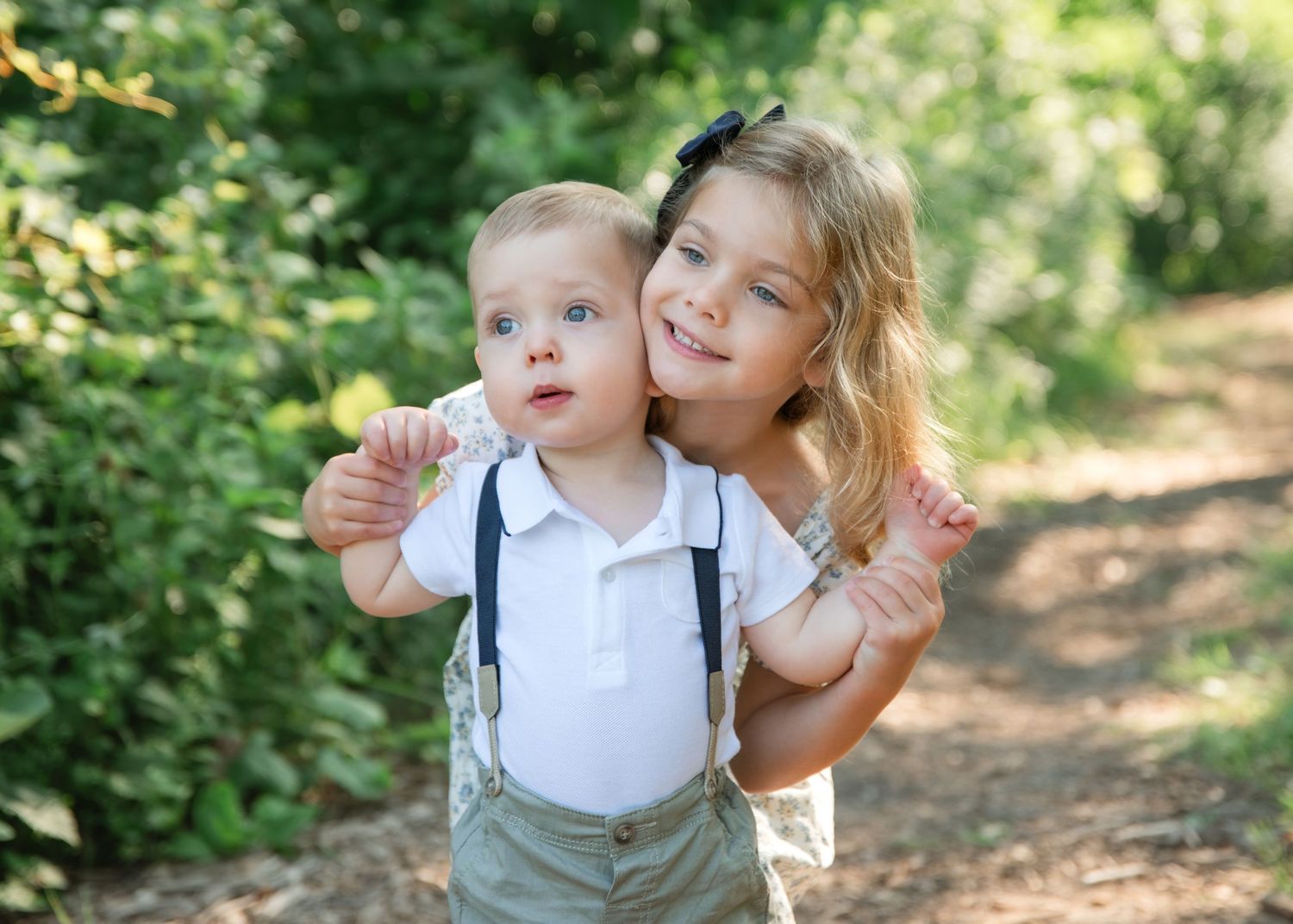 Older girl hugging baby boy in suspenders outdoors on a sunny garden path surrounded by lush green foliage.
