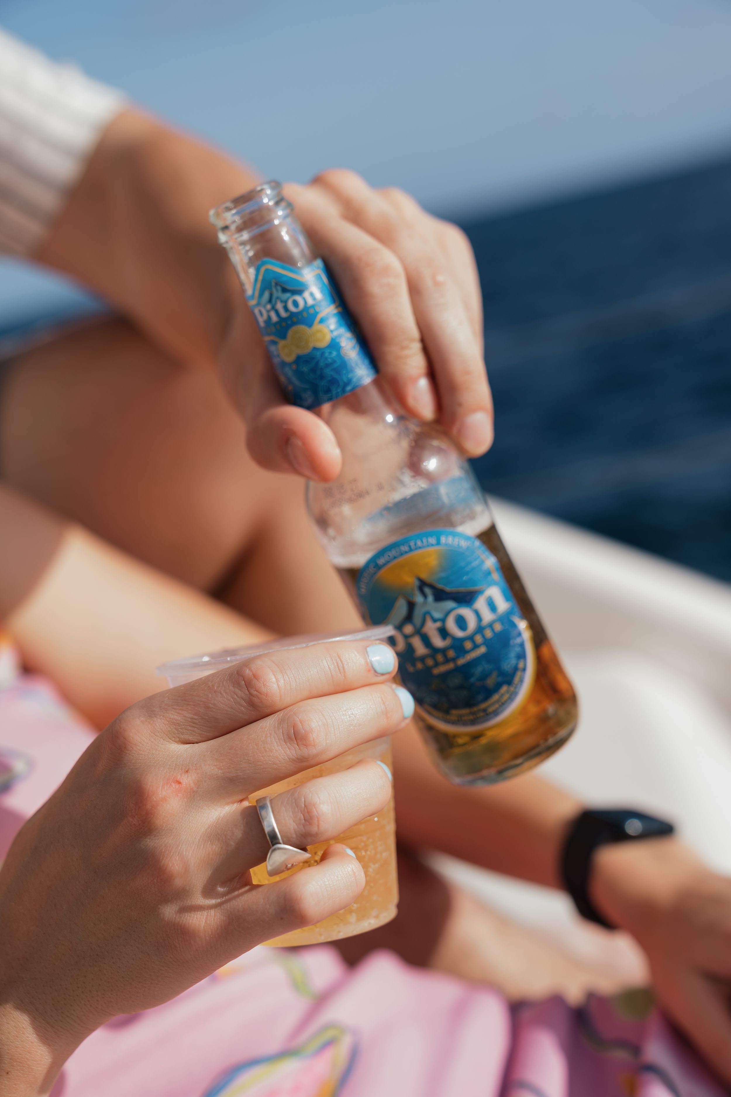 Two people clinking Piton beer bottles on a sunny boat deck with ocean views in the background.