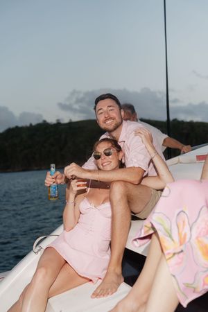 Couple smiling on a boat at sunset, holding drinks, wearing pink outfits, with tropical island in background.