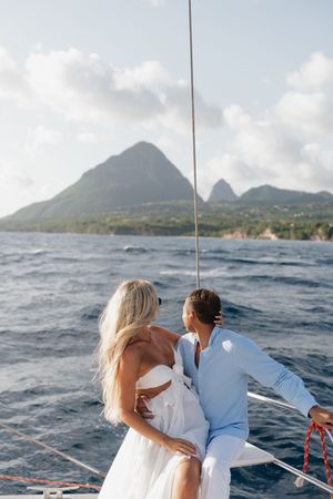 Couple in white attire embracing on a sailboat with stunning tropical mountain views in the background.
