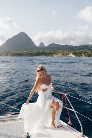 Blonde woman in white dress on yacht bow with scenic Piton mountains of St. Lucia in background.