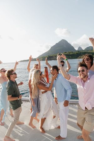 A joyful group celebrates on a yacht deck with stunning mountain views, dancing and raising hands in the sunny Caribbean.