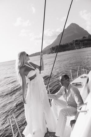 Black and white photo of a couple on a sailboat, woman in white dress holding rigging, mountain backdrop.