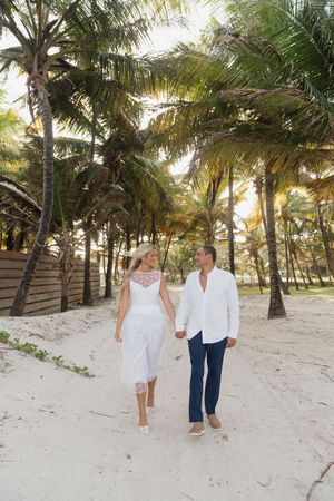 Bride and groom walk hand in hand along sandy path through lush tropical palm tree grove.