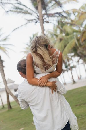 Groom lifts bride in lace wedding dress outdoors near tropical palm trees on wedding day.