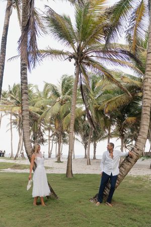 Couple in white outfits posing among tropical palm trees on a sandy beach, perfect for destination wedding inspiration.