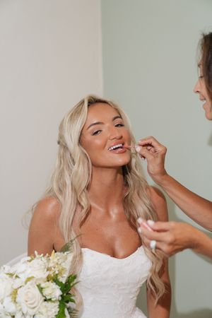 Bride with blonde hair getting lipstick applied by makeup artist while holding white bouquet in strapless wedding gown.