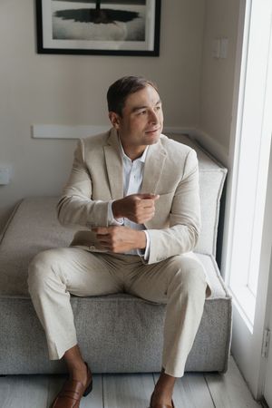 A man in a beige linen suit sits on a sofa, gazing out a window in a bright modern room.