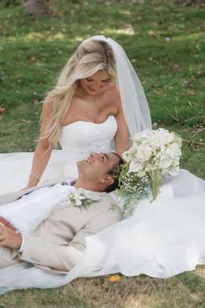 Bride in white strapless gown with veil smiles at groom lying on grass holding white rose bouquet.