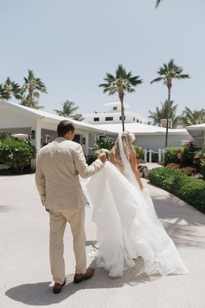 Bride and groom walking hand in hand toward a tropical resort, with palm trees and white buildings under a sunny sky.