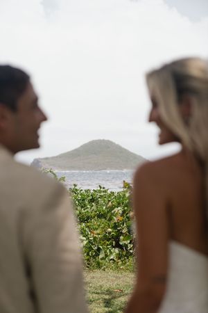 A bride and groom face each other with a tropical island and ocean view in the background.