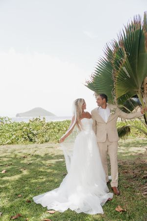 Bride and groom share romantic moment near tropical palm tree with ocean view backdrop.
