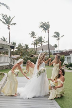 Bride surrounded by bridesmaids in yellow dresses adjusting her gown on a tropical venue deck with palm trees.