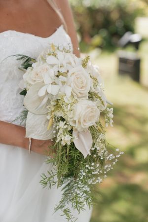 Bride holding a cascading white wedding bouquet with roses, orchids, baby's breath, and greenery in an outdoor setting.