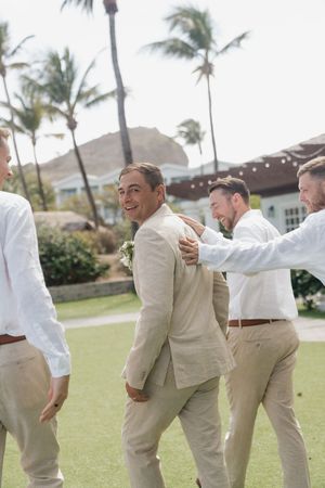 Groom in tan suit laughs with groomsmen in white shirts on tropical resort lawn with palm trees.