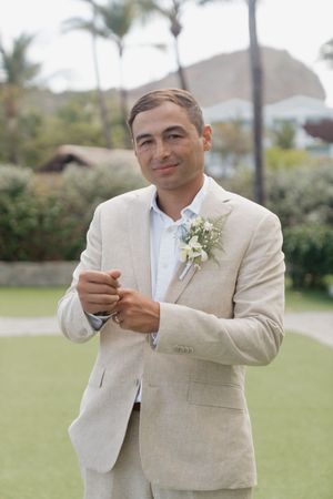 Groom in beige linen suit with boutonniere smiling outdoors at tropical wedding venue with palm trees.