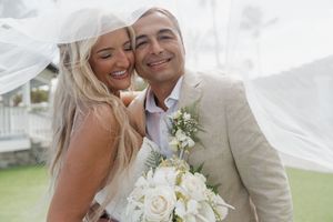 Bride in white veil embraces groom in beige suit holding white rose bouquet at outdoor wedding ceremony.