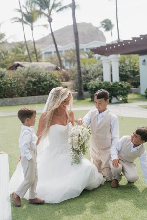Smiling bride in white gown kneels with three young ring bearers in beige suits outdoors.