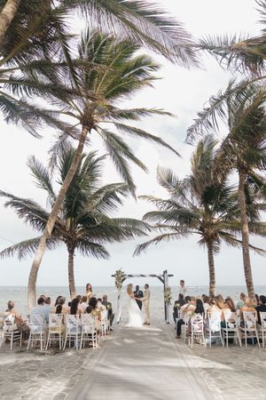 Beach wedding ceremony under palm trees with guests seated on white chairs facing couple at floral arch by the ocean.