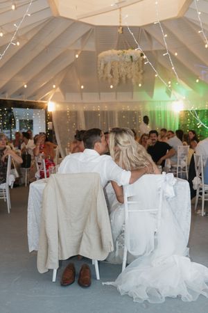 Bride and groom sit at wedding reception table under fairy lights in elegant white tent venue.