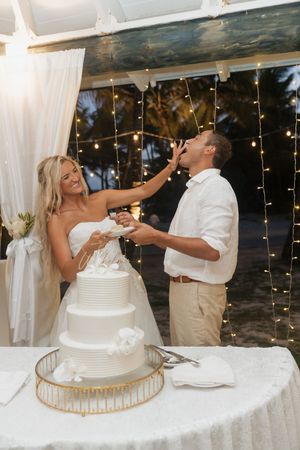 Bride feeding wedding cake to groom at elegant tropical reception with string lights and white tiered cake.