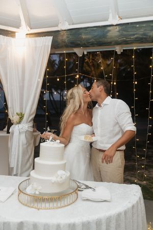 Bride and groom share a kiss while cutting their white tiered wedding cake under warm string lights.