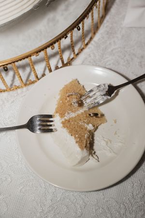 A partially eaten slice of white frosted cake on a plate with two forks at an elegant wedding reception.