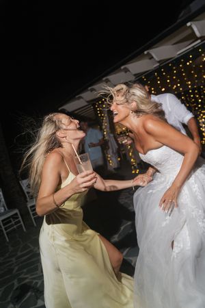 Bride in white gown and bridesmaid in yellow dress dancing joyfully at outdoor evening reception.