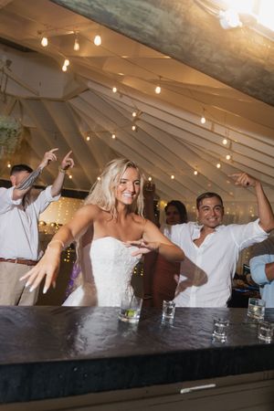 Joyful bride in strapless gown dancing with guests at an outdoor tented wedding reception with string lights.