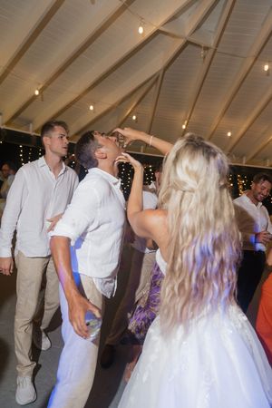 Bride and groom dancing joyfully at wedding reception under string lights in a tent with guests celebrating.