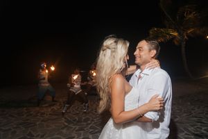 Bride and groom share a romantic embrace on a beach at night with fire performers in the background.