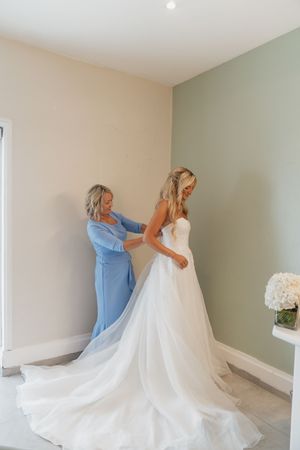 A bride in a white wedding gown gets her dress fastened by a woman in a blue dress in a bright room.