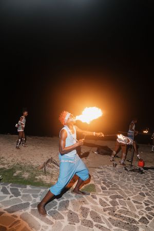 Fire breather performing at night on a sandy beach, exhaling a large flame during a cultural fire show.