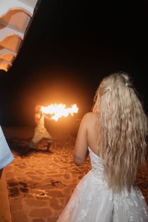 Bride watches fire dancer perform on sandy beach during nighttime tropical wedding celebration.