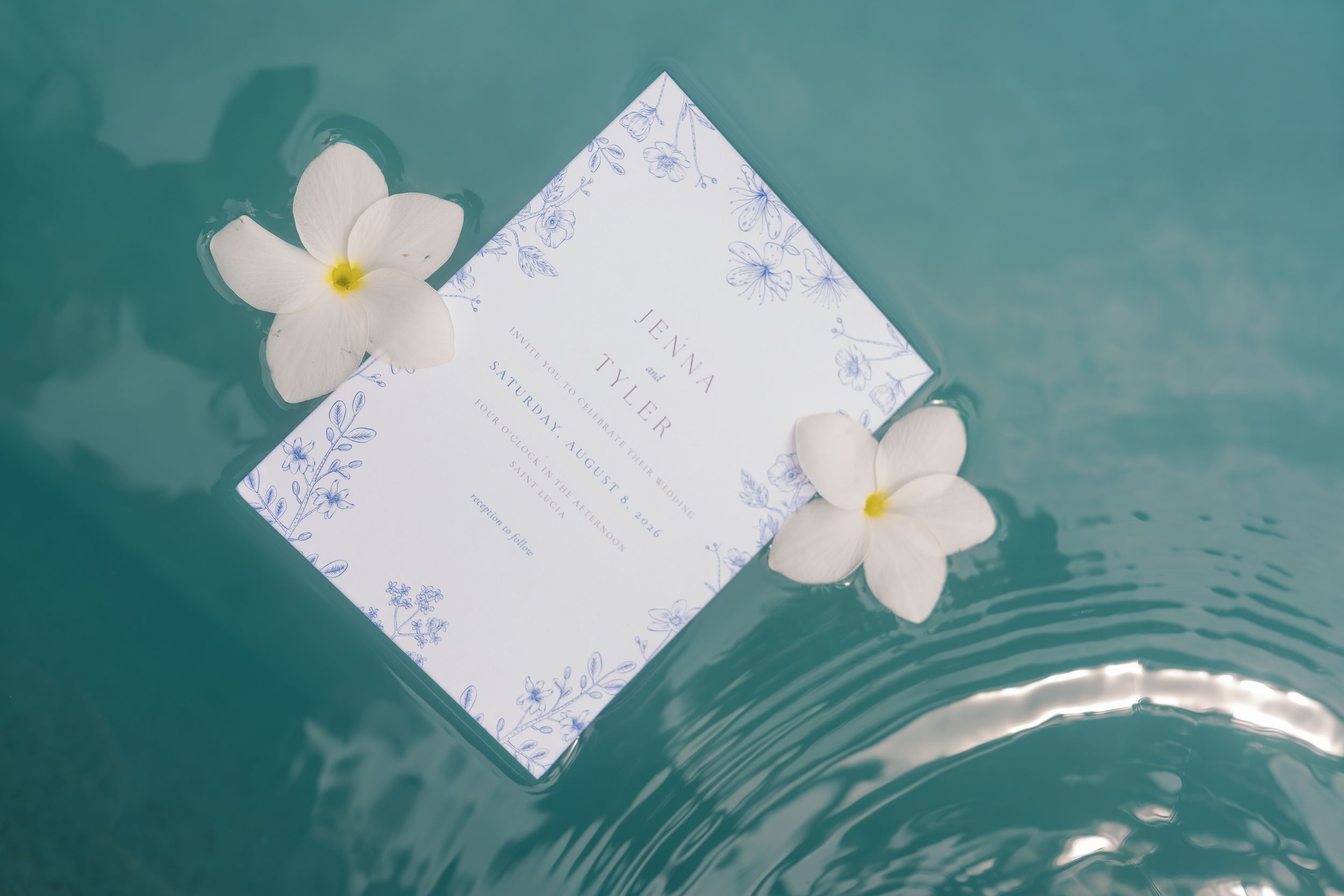 A floral wedding invitation card floats on turquoise water, adorned with two white plumeria flowers.