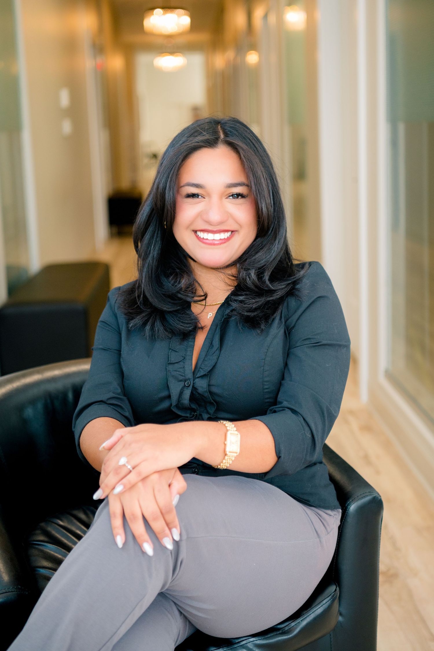 Smiling professional woman in black blouse and gray pants seated in leather chair in modern office hallway.
