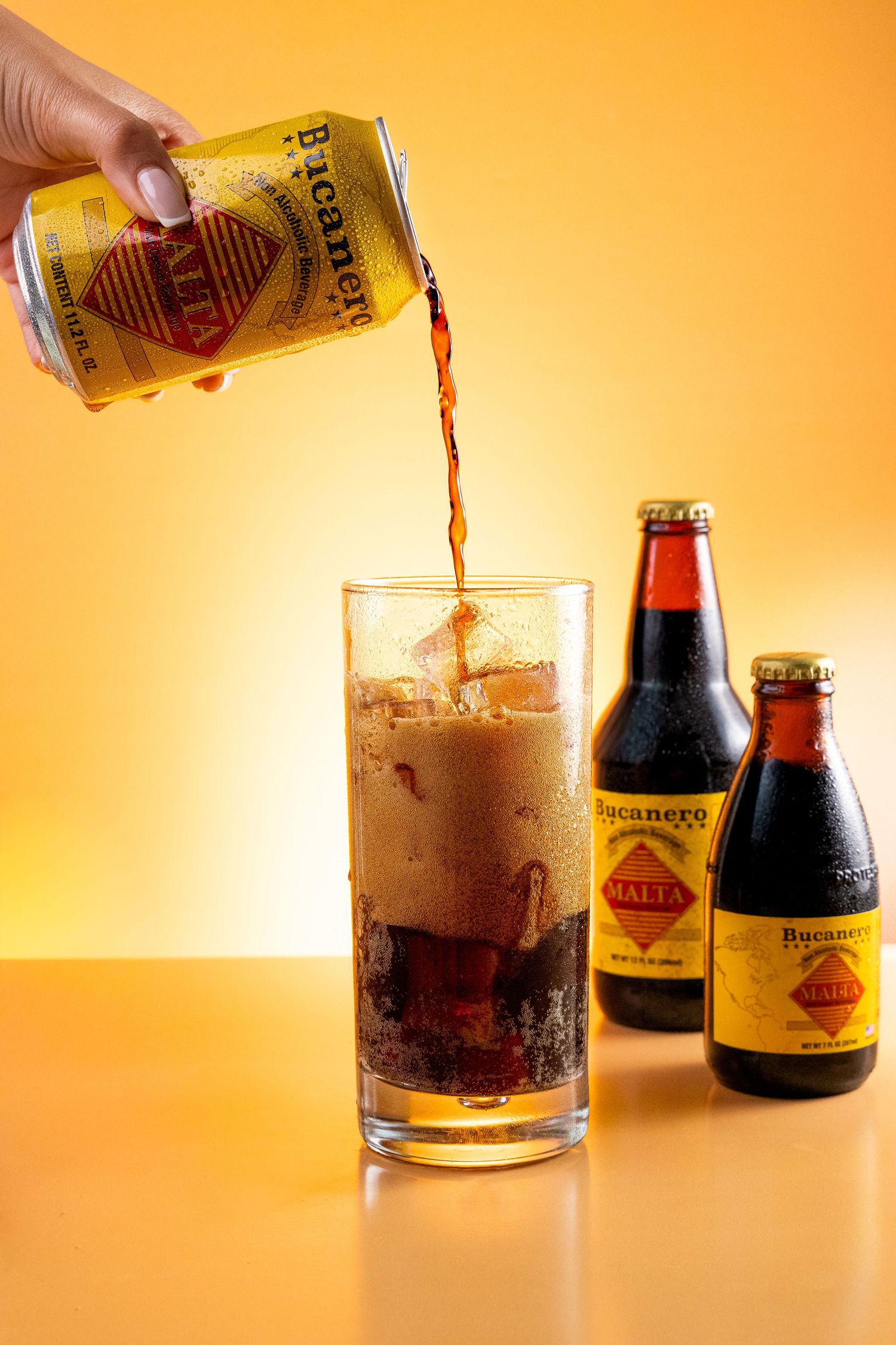 Bucanero Malta being poured from a can into an ice-filled glass, with two bottles on an orange background.