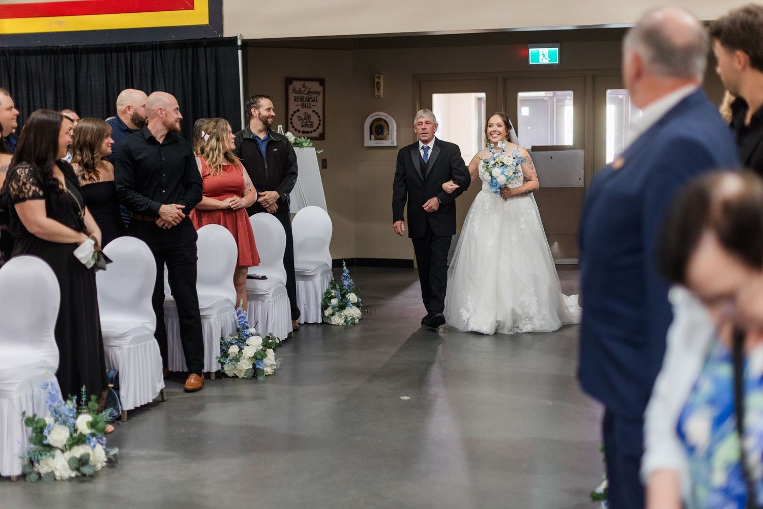 Bride walks down the aisle with her father at an indoor wedding ceremony with guests seated on white chairs.